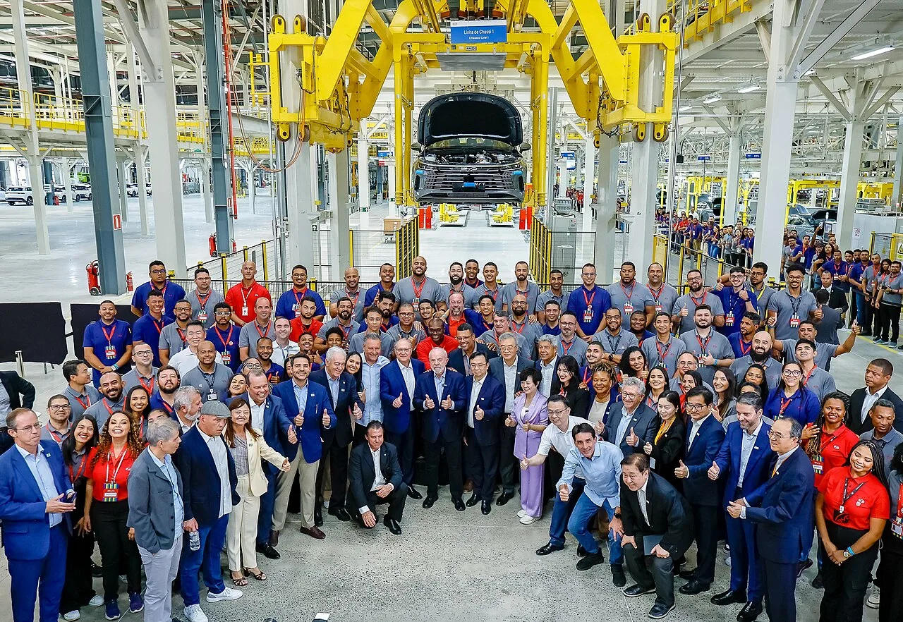 A large group of workers standing on a car assembly line under a car body hanging from an overhead carrier