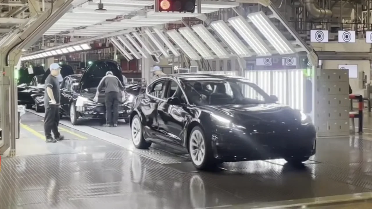 A black Tesla sedan at the end of an assembly line under bright inspection lights, with workers checking cars behind it