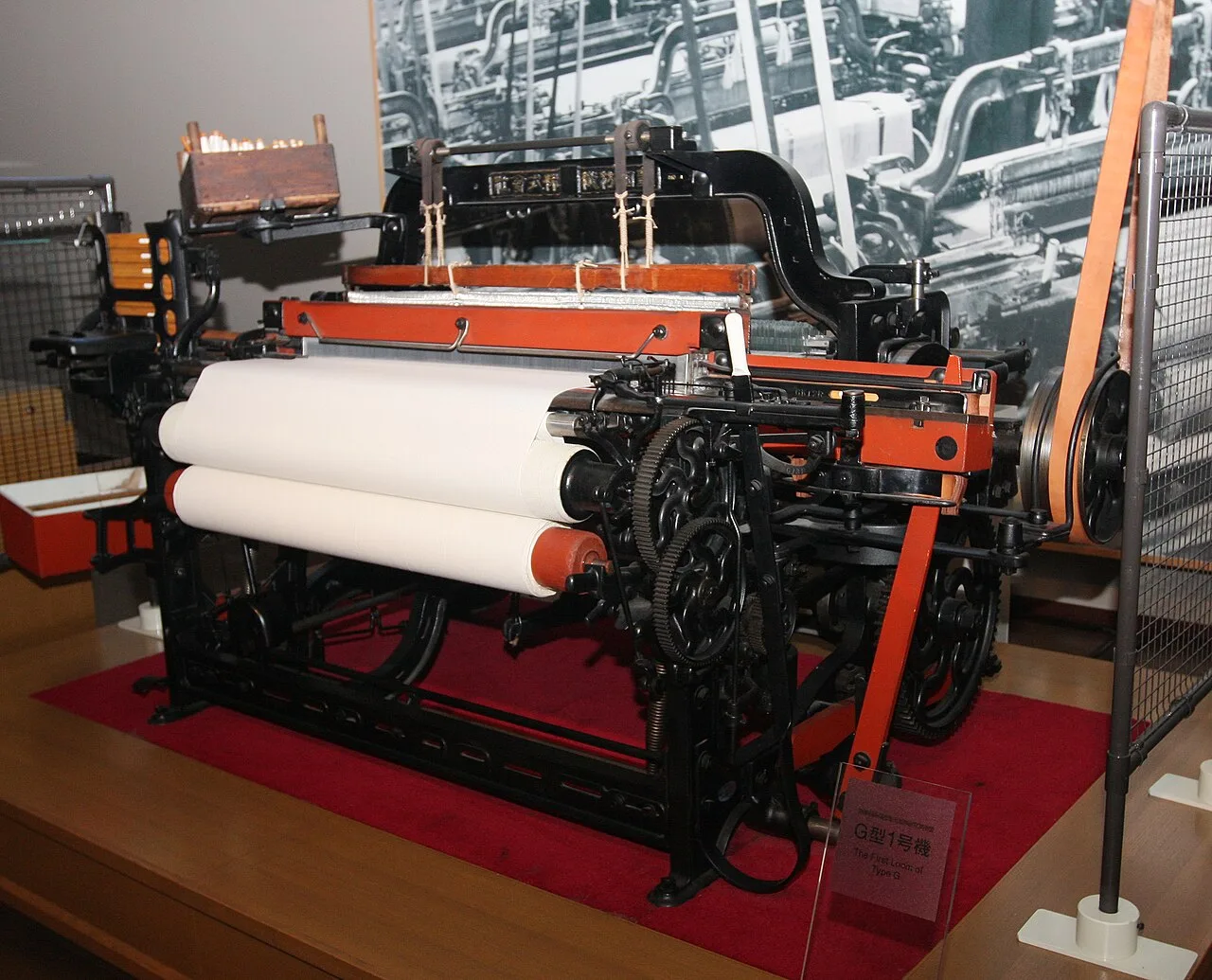 A black and orange cast-iron loom with white cloth rolls, on display in a museum