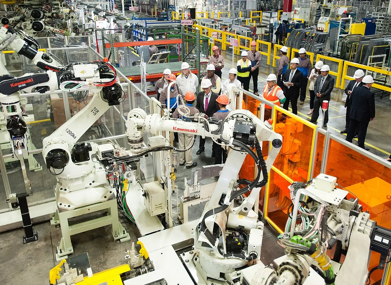 White welding robots behind orange safety screens, with a group in hard hats walking past the cell