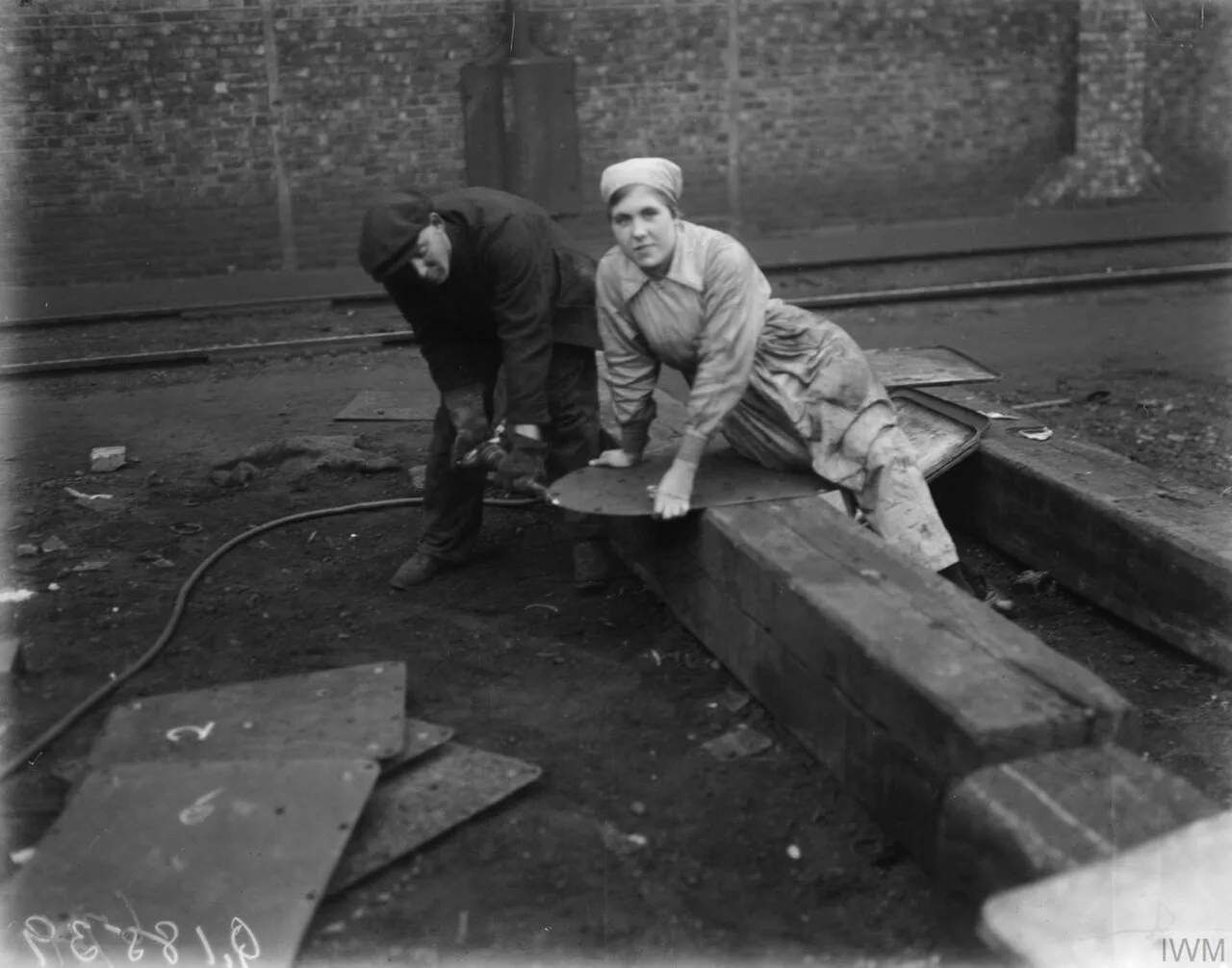 A man and a woman riveting a steel plate on a wooden block in a shipyard