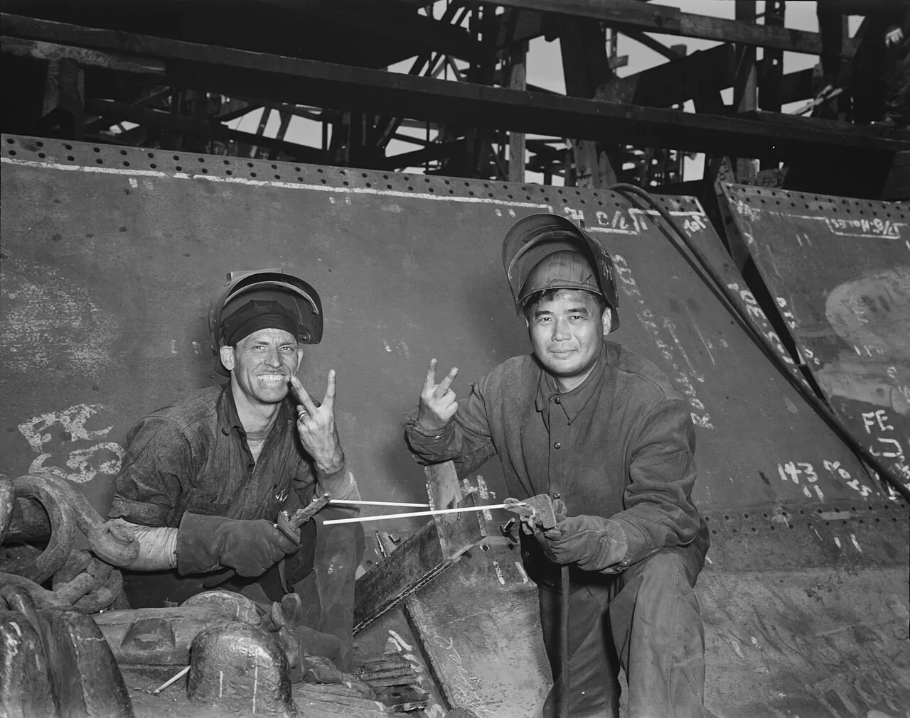 Two welders in protective hoods sitting against the steel hull of a ship under construction